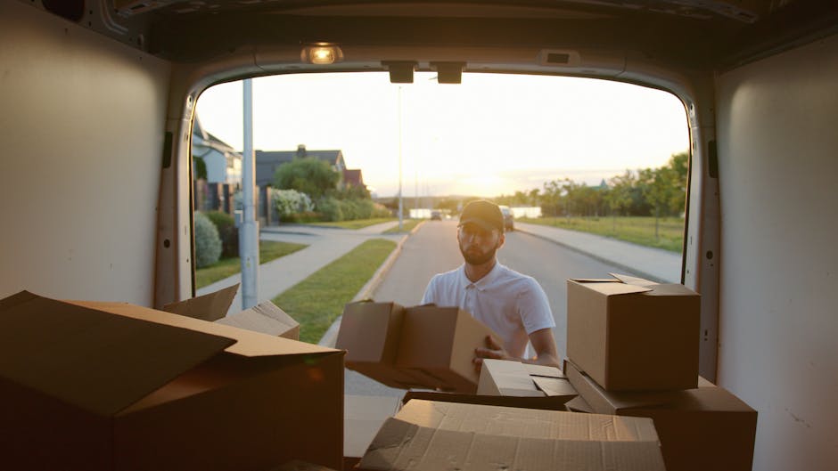A man wearing a white t-shirt, jeans, and a black cap is loading large cardboard boxes into the side of a white commercial van parked on a residential street during daytime. The van, positioned parall