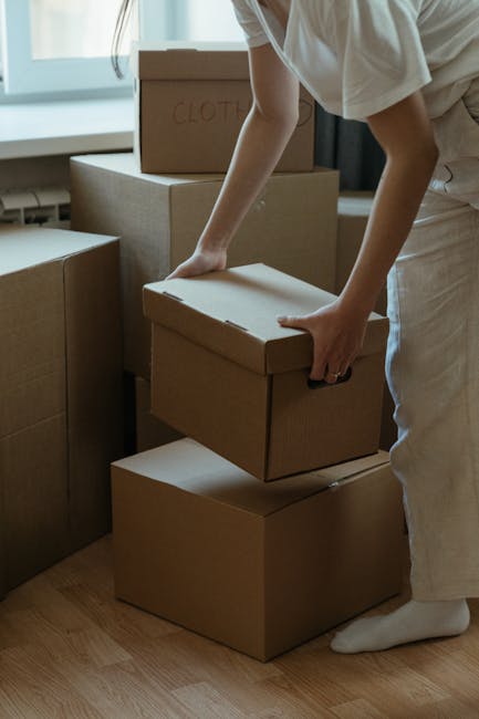 A person wearing light-colored clothing is seen stacking several cardboard boxes in a room with natural daylight coming through a window. The boxes are of varying sizes, with one labeled 'CLOTH' written in red marker, indicating packing for a home relocation. The individual is holding a medium-sized box, preparing it for transportation, possibly as part of furniture transport or packing and moving activities coordinated by Finchley Movers. The environment appears to be indoors, near a doorway or window, with additional boxes placed nearby on a wooden floor. Visible equipment includes the cardboard boxes themselves, and the process reflects the loading phase of a house removal, where careful handling and organization of goods are essential for a smooth move.