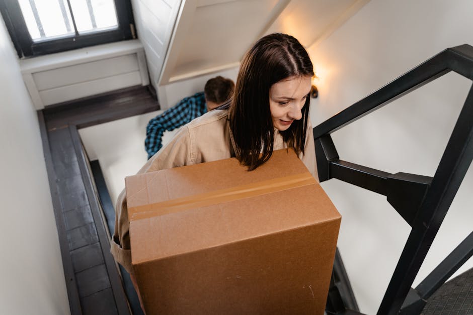 A woman with shoulder-length dark hair, wearing a beige jacket, is carrying a large cardboard box up a narrow staircase inside a home. Behind her, a person in a blue checkered shirt is visible, assisting with the move. The staircase has black metal railings, white painted walls, and hardwood flooring. The cardboard box appears to be securely sealed with packing tape, and the woman is carefully navigating the steps, focusing on her task during the home relocation process. The environment is well-lit with natural light coming from a window on the upper landing, and the scene illustrates packing and furniture transport activities typical of residential moves. The overall setting emphasizes internal movement logistics, consistent with professional removals services, such as those provided by Finchley Movers.