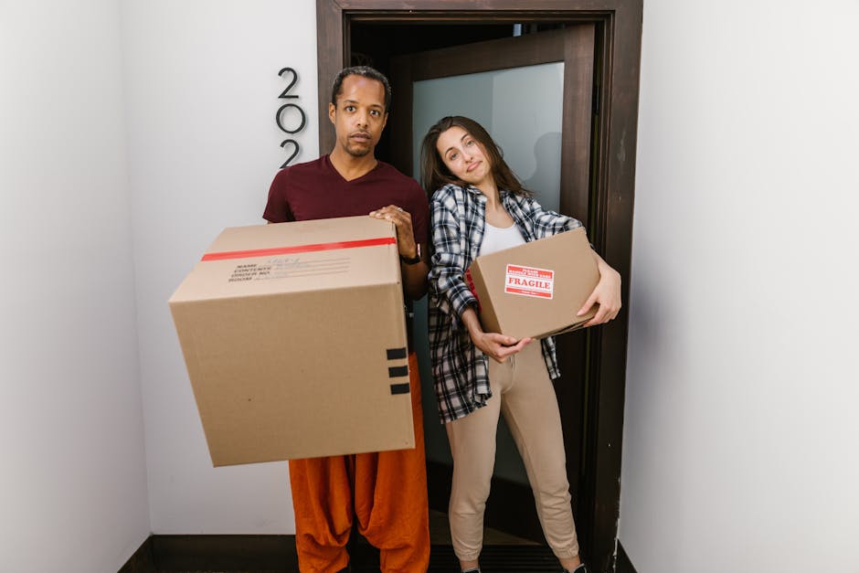 A person wearing light-colored clothing is seen stacking several cardboard boxes in a room with natural daylight coming through a window. The boxes are of varying sizes, with one labeled 'CLOTH' written in red marker, indicating packing for a home relocation. The individual is holding a medium-sized box, preparing it for transportation, possibly as part of furniture transport or packing and moving activities coordinated by Finchley Movers. The environment appears to be indoors, near a doorway or window, with additional boxes placed nearby on a wooden floor. Visible equipment includes the cardboard boxes themselves, and the process reflects the loading phase of a house removal, where careful handling and organization of goods are essential for a smooth move.
