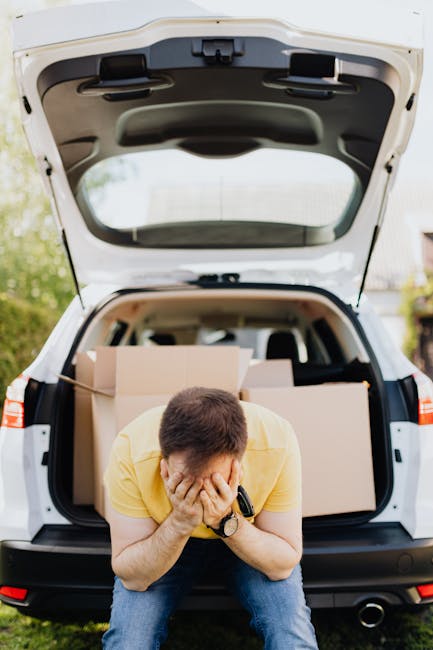 A man wearing a yellow shirt and blue jeans is sitting on the ground in front of an open car trunk, covering his face with both hands. The trunk is filled with cardboard boxes used for packing and moving household items, with some boxes appearing to be securely sealed. The vehicle is a white hatchback or small SUV, parked outdoors on a paved or grassy area, with additional boxes visible inside the car. The scene depicts the physical and emotional aspects of home relocation, involving furniture transport and packing preparations, with a focus on the loading process as part of an organized removals service provided by Finchley Movers, supporting moves like those described in the Finchley Central removals guide for Ballards Lane moves.