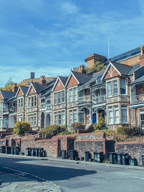 A row of Victorian-style terraced houses on a residential street, featuring brick facades, decorative ironwork on balconies and porches, and large bay windows, with some greenery and small trees in front gardens. The street level includes a pavement with multiple black wheelie bins lined up along the curb. The sky is blue with some light clouds, and a building with a glass rooftop is visible in the background. This setting reflects a typical London neighbourhood suitable for house removals and home relocation services, such as those offered by Finchley Movers, facilitating furniture transport and packing logistics for moving house in North Finchley N12 near Artsdepot.
