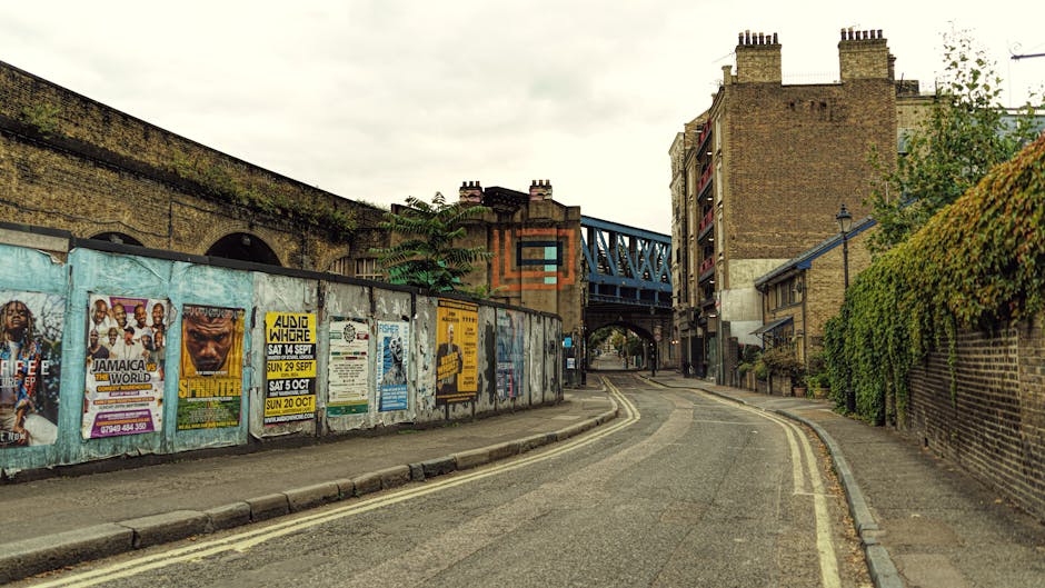 A narrow urban street with a curved section leading towards a railway bridge overhead in North Finchley N12. The road is flanked by brick and concrete buildings, with a brick wall covered in green foliage on the right side and a weathered blue and white advertising poster wall on the left. The street surface features double yellow lines along the curb, indicating parking restrictions, and the area appears to be in a quiet residential or mixed-use neighbourhood. The scene is captured during daylight, with overcast skies providing diffuse lighting. This setting is typical for house removals and moving services, with potential access points for loading and unloading furniture near the railway bridge, supporting logistical operations by Finchley Movers for home relocation or furniture transport within the area.